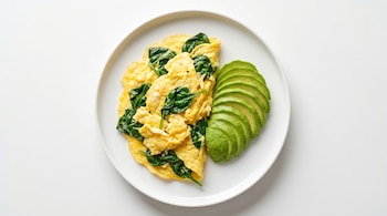 Vista aérea de huevos revueltos mezclados con espinacas marchitas en un plato blanco, con aguacate en rebanadas ordenadas a un lado.