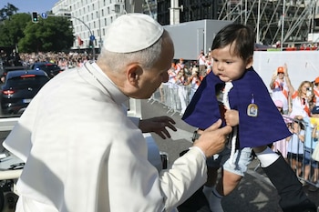 Un pontífice con vestimenta blanca extiende las manos hacia un niño con capa morada sostenido en brazos junto a un vehículo en la calle