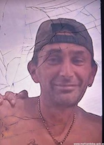 Fotografía de un hombre con gorra hacia atrás y cadena en el cuello, vista a través de un cristal con grietas pronunciadas