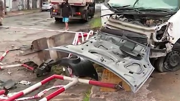 Una camioneta blanca severamente dañada con el capó abierto y partes destrozadas yace en una calle mojada, rodeada de escombros y cinta de seguridad