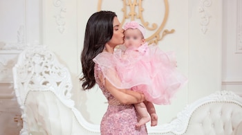 Ely Yutronic, con vestido rosa brillante, besa la frente de su hija Luna, quien viste un tutú rosa y diadema de flores. El rostro de la bebé está borroso. Fondo blanco con decoraciones.