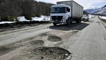 Un camión blanco transita por una carretera de asfalto con grandes baches, rodeada de nieve y montañas al fondo