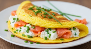 Omelette amarillo doblado con relleno de queso crema blanco, tiras rosadas de salmón ahumado y cebollín verde picado en un plato blanco.