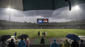 Estadio de fútbol con campo mojado por lluvia intensa. Marcador electrónico muestra icono de lluvia y alerta 17:00. Personas con paraguas y ponchos.