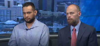 Dos hombres sentados con micrófonos. Uno con camisa de cuadros y barba, el otro con traje y corbata. Fondo digital muestra un paisaje urbano