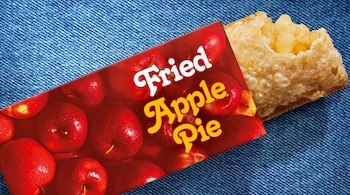 Un pastel de manzana frito dorado, con el relleno de manzana expuesto, sobresale de un envoltorio rojo con diseño de manzanas y texto