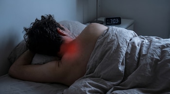Un hombre duerme de lado en la cama con una manta gris. La nuca y parte de la espalda alta se ven con un color rojizo. Un reloj digital marca las 7:03.