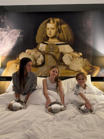 Una mujer y dos niños sentados en una cama con sábanas blancas, con tres cloches plateados. Detrás, un cuadro grande de una niña