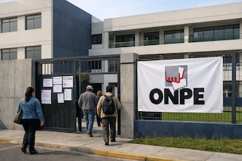 Fachada de un colegio moderno en tonos gris y blanco con una reja negra. Una pancarta blanca con el logo de ONPE y personas entrando al local de votación.