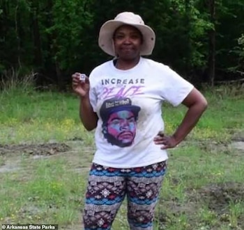 Una mujer sonriente con sombrero claro y camiseta estampada sostiene un pequeño diamante blanco en su mano derecha, posando en un campo verde y terroso