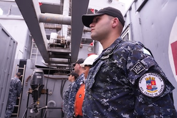 Varios militares con uniforme de camuflaje azul y gorra en la cubierta de un barco junto a una grúa y un motor fuera de borda