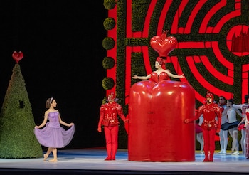 Bailarina con vestido lila, una mujer con traje rojo que simula un corazón, dos guardias rojos, otros bailarines y escenografía de jardín con fondo rojo y verde