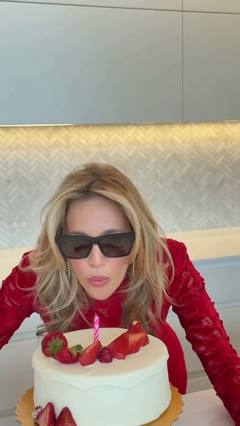 Luisana Lopilato, con gafas de sol y vestido rojo, se inclina para soplar una vela rosa en una torta blanca decorada con fresas en una cocina moderna