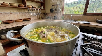 Olla grande de metal hirviendo con caldo de costilla, papas y cebolla larga, sobre una estufa en una cocina con azulejos y utensilios.