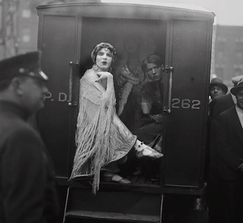 Fotografía en blanco y negro de Mae West, vestida con un chal con flecos y tocado, sentada dentro de un furgón policial, con un oficial a la izquierda