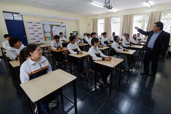 En un aula, un profesor señala mientras varios estudiantes jóvenes uniformados con chaquetas blancas y azules atienden desde sus pupitres individuales