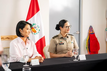 Dos mujeres sentadas frente a una mesa con copas de agua y letreros de identificación, con una bandera de Perú al fondo