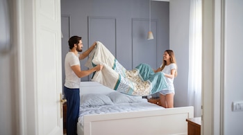 Un hombre y una mujer sonrientes, de pie a cada lado de una cama blanca, extienden un edredón estampado con motivos verdes, dorados y blancos.