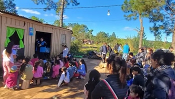 Un grupo de personas y niños sentados en el suelo frente a una edificación de madera con árboles al fondo