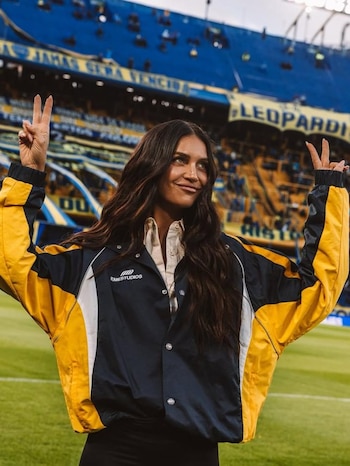 Zaira Nara sonríe y hace el signo de la paz con ambas manos. Viste una chaqueta azul y amarilla. Está en el césped de un estadio de fútbol con gradas de fondo