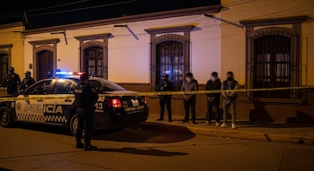 Imagen nocturna en Puebla: Una casa con cinta "No pasar", una patrulla policial estacionada y tres hombres esposados siendo custodiados por policías con cubrebocas.
