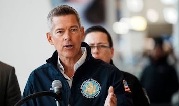 Sean Duffy, con una chaqueta azul del Departamento de Transporte de EE. UU. y la bandera estadounidense en la manga, habla en un micrófono plateado. Fondo interior borroso