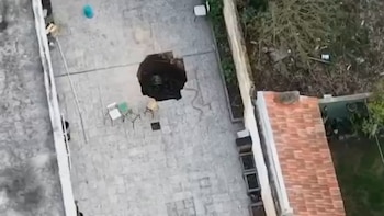Vista aérea de un patio pavimentado con un socavón oscuro en el centro, rodeado por sillas, jardineras y un tejado de tejas
