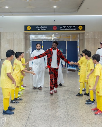 Un hombre vestido con un traje deportivo rojo y negro sonríe mientras camina entre dos filas de niños que visten uniformes de fútbol amarillos en un aeropuerto
