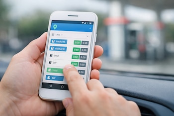 Mano sosteniendo un smartphone blanco con la aplicación Facilito de Osinergmin abierta, mostrando precios de gasohol, diésel y GLP en Lima. Fondo de estación de servicio.