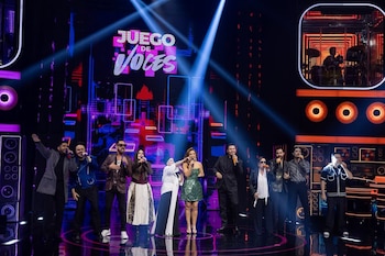 Grupo de cantantes masculinos y femeninos en un escenario oscuro con luces brillantes azules y moradas. El logo "Juego de Voces" está al fondo. Un baterista se ve en pantalla superior