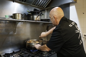 Hombre calvo en chaqueta de chef negra remueve alimentos en una olla sobre fogón de gas. Estantes metálicos con ollas y sartenes