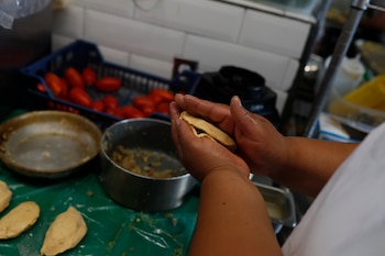 Una mujer sonriente con gorro y delantal blanco con un logo de comida, sostiene una bandeja negra con tres platos de comida. Al fondo, personas y pancartas