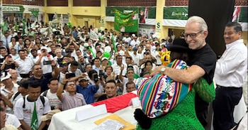 Ricardo Belmont, un hombre mayor con gafas y camisa negra, abraza a una mujer vestida con una manta andina colorida en un mitin político abarrotado de personas