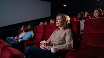 Una mujer sonriente en un cine oscuro, sosteniendo palomitas y una bebida, mira la pantalla junto a otros espectadores en asientos rojos de terciopelo.