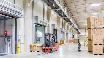 Interior de un gran almacén o fábrica con múltiples muelles de carga, un montacargas rojo, un trabajador y pilas de cajas de cartón sobre palés