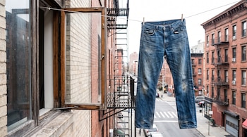 Pantalón vaquero azul colgado con pinzas en un tendedero que sale de una ventana abierta, frente a edificios de ladrillo, escaleras de emergencia y una calle.