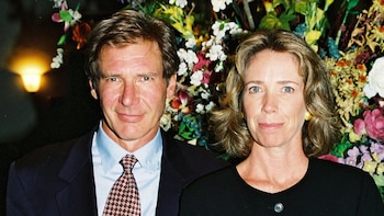 Harrison Ford con traje oscuro y corbata junto a Melissa Mathison vestida de negro con un arreglo floral de fondo