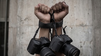 Primer plano de dos manos alzadas, una de piel clara y otra oscura, atadas por las correas de dos cámaras profesionales negras, frente a un muro de cemento.