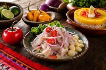 Un plato de ceviche de pescado con cebolla morada y choclo junto a una causa limeña, ajíes y limones sobre una mesa de madera.