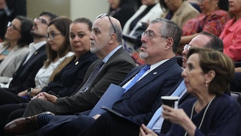 Vista lateral de una fila de seis personas adultas sentadas, la mayoría usa gafas y ropa formal. Un hombre sujeta una carpeta, una mujer una taza