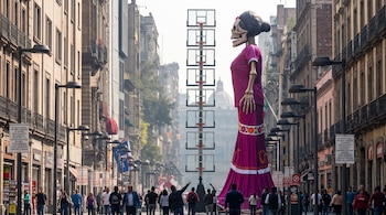 Estatua gigante de la Catrina junto a una torre de diez canastas de baloncesto en una avenida peatonal flanqueada por edificios y transeúntes.