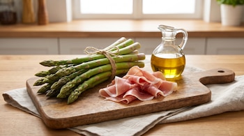 Espárragos verdes atados con cuerda, fetas de jamón crudo y una jarra de vidrio con aceite de oliva sobre una tabla de cortar de madera.