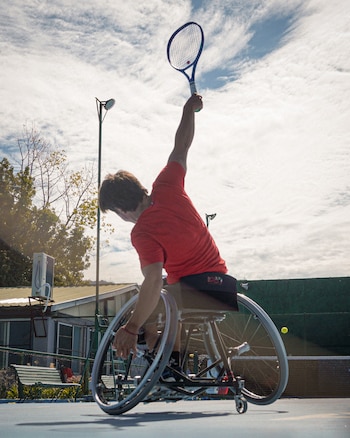 Gonzalo Lazarte en silla de ruedas, vestido con una camiseta roja, golpea una pelota de tenis con una raqueta en una cancha azul. El cielo está parcialmente nublado