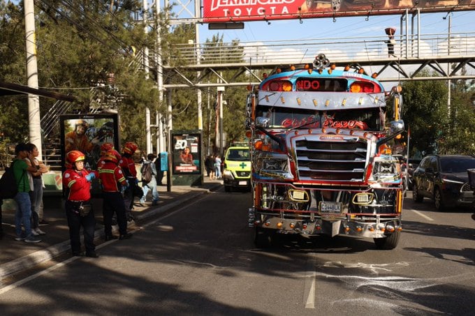 Un grupo de socorristas permanece en la acera mientras un autobús decorado transita por la calle junto a un puente peatonal. (Bomberos Municipales de Guatemala)