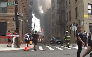Calle en el Bronx con edificios, humo negro, fuego, camión de bomberos, mangueras, policía, bomberos, civiles y un coche estacionado
