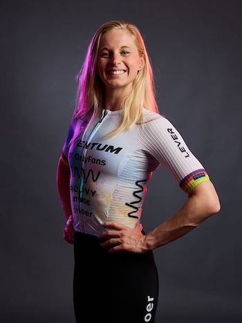 Retrato de mujer ciclista rubia sonriendo a cámara, vestida con maillot blanco con logotipos y pantalón negro, manos en las caderas. Luz rosa a la izquierda