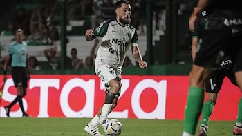 Primer plano de Jonatan Gomez con barba, vistiendo un uniforme de fútbol blanco y verde, driblando el balón en un campo verde