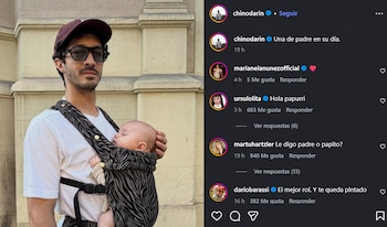 Un hombre con gorra y gafas de sol sostiene a un bebé dormido en un portabebés con estampado de cebra, frente a una pared de piedra