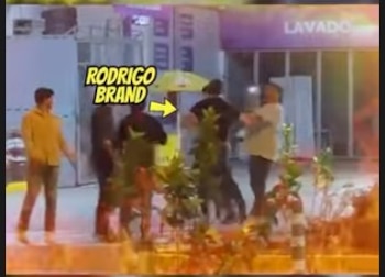 Un hombre, identificado como Rodrigo Brand por una flecha amarilla, de espaldas con una chaqueta oscura, en un área exterior con otras personas. Al fondo, un letrero de 'LAVADO'