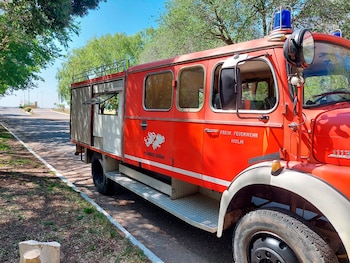 Camión de bomberos rojo con cabina blanca, luces de emergencia azules, texto en el lateral y calcomanía de Malvinas, estacionado en carretera arbolada
